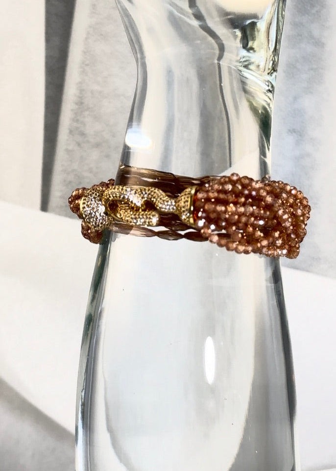 Gold and brown beaded bracelet, crystal hook clasp, on a clear glass display with a blurred white background